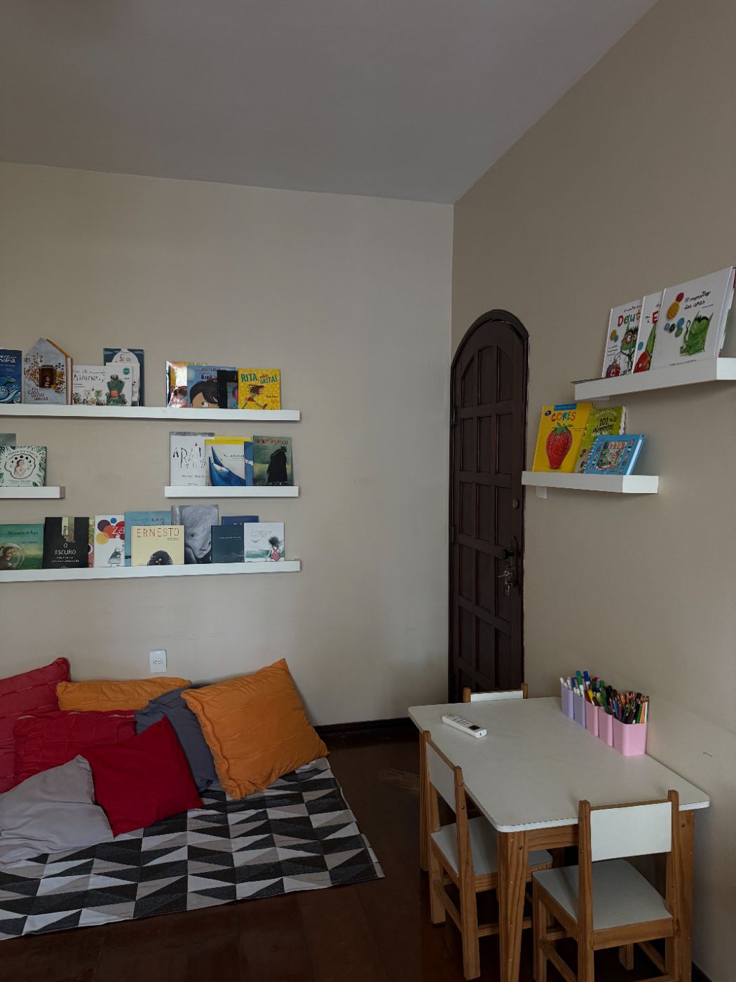 Parede de prateleiras com livros infantis, tapete com almofadas e mesa de atividades